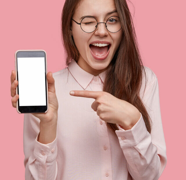cropped-look-at-this-cell-phone-pleased-happy-woman-blinks-eyes-points-with-index-finger-at-blank-screen-shows-modern-device.jpg
